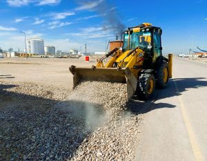 A yellow excavator unloading pebbles on a road at an industrial construction site under a clear blue sky.