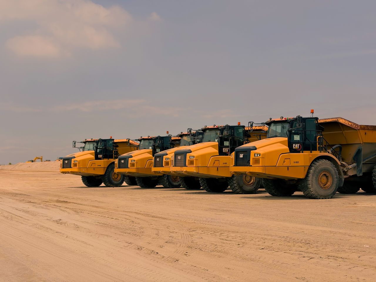 about-02 Row of heavy-duty dump trucks parked on a sandy construction site under a clear sky.