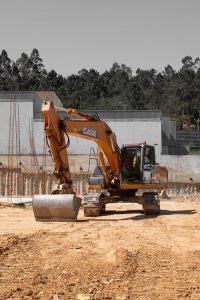 An excavator at a construction site against a forested backdrop under a clear sky.