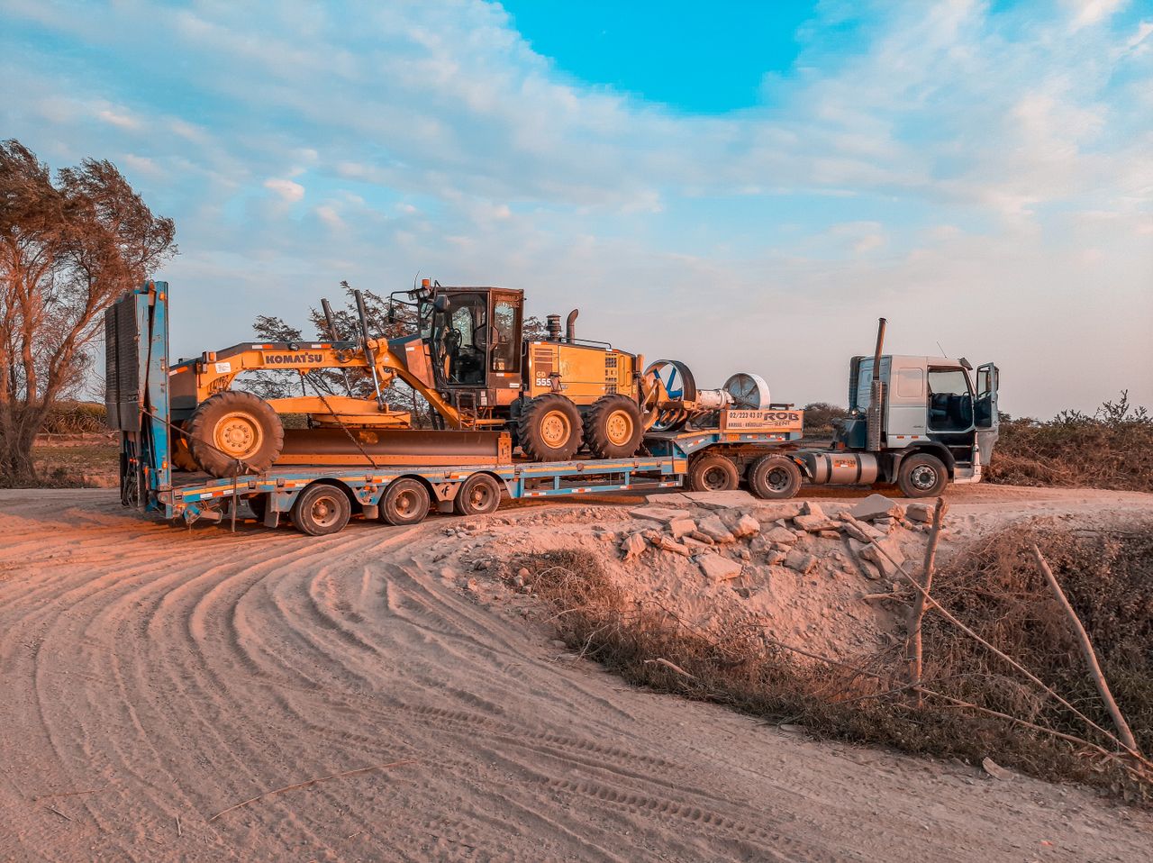 why-choose-us A cargo truck transporting heavy machinery on a rugged dirt road under a clear sky.